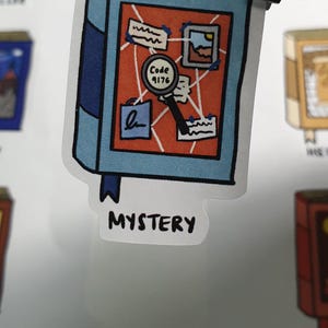 Detailed close up shot of a Mystery book sticker. The sticker is Blue and Orange. The sticker is held by a tweezer against a blurred background of the sticker sheet.