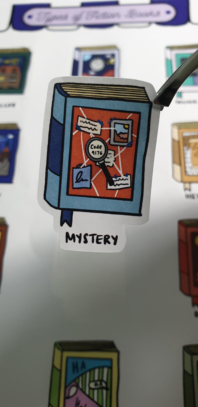 Detailed close up shot of a Mystery book sticker. The sticker is Blue and Orange. The sticker is held by a tweezer against a blurred background of the sticker sheet.