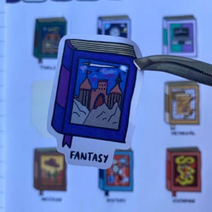 Detailed close up shot of a Fantasy book sticker. The sticker is Purple and Blue and features a beautiful castle. The sticker is held by a tweezer against a blurred background of the sticker sheet.