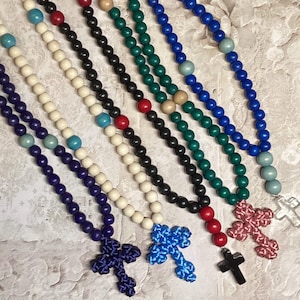 May include: A collection of colorful rosaries with wooden beads and cross pendants. The rosaries feature various colors, including blue, black, white, green, and red. The crosses are made of different materials and colors.
