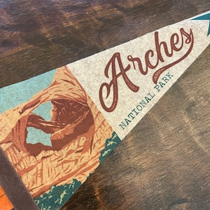 May include: A brown and tan pennant with the text "Arches National Park" and a picture of a natural rock arch.