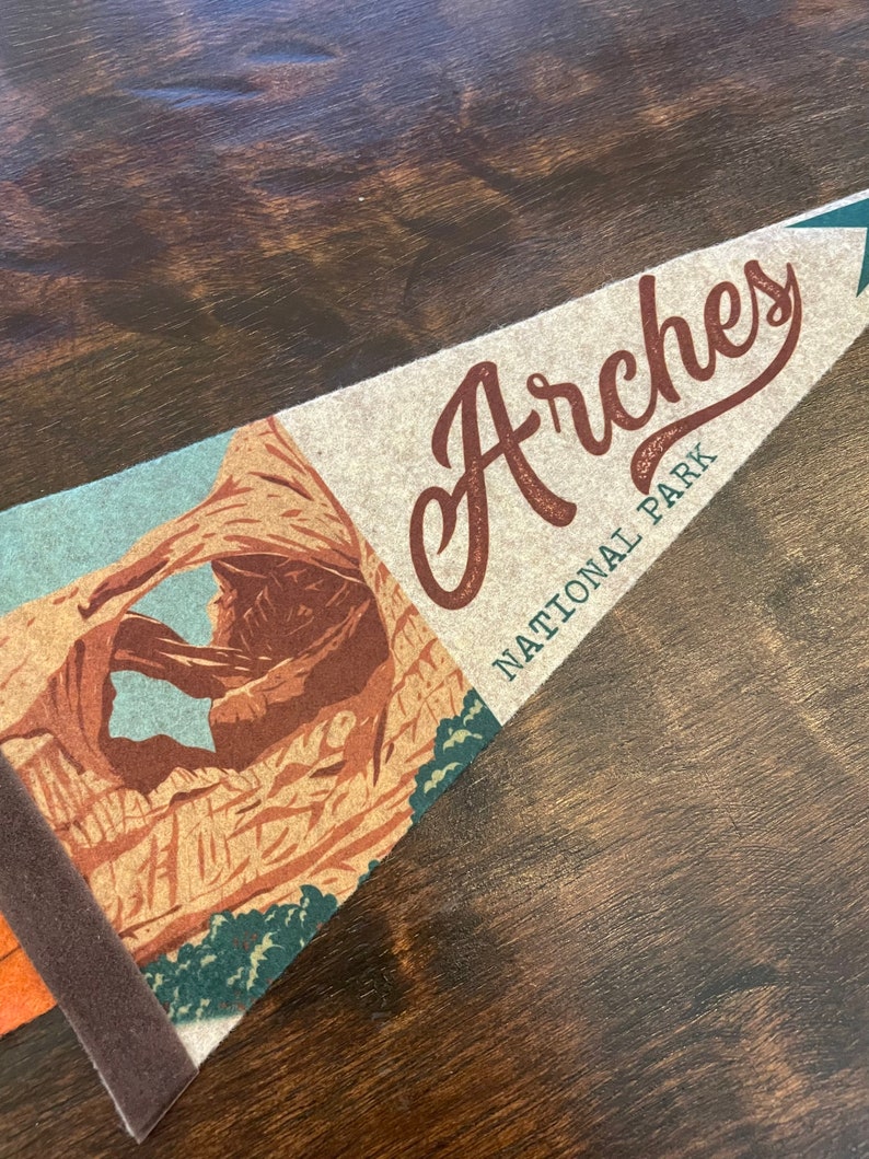 May include: A brown and tan pennant with the text "Arches National Park" and a picture of a natural rock arch.
