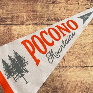 May include: A white pennant with an orange border and the text "POCONO Mountains PA" in orange and gray lettering. The pennant features a gray silhouette of two pine trees.