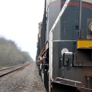 Puede incluir: Primer plano de una locomotora de tren verde oscuro y gris con una franja amarilla y un faro redondo. El tren está en un conjunto de vías de ferrocarril.