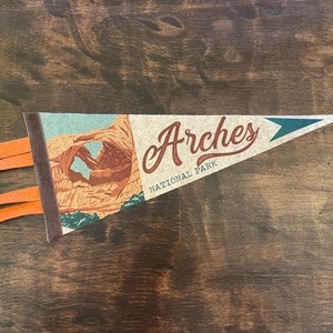 May include: A brown and tan pennant with a picture of a rock formation and the text "Arches National Park".