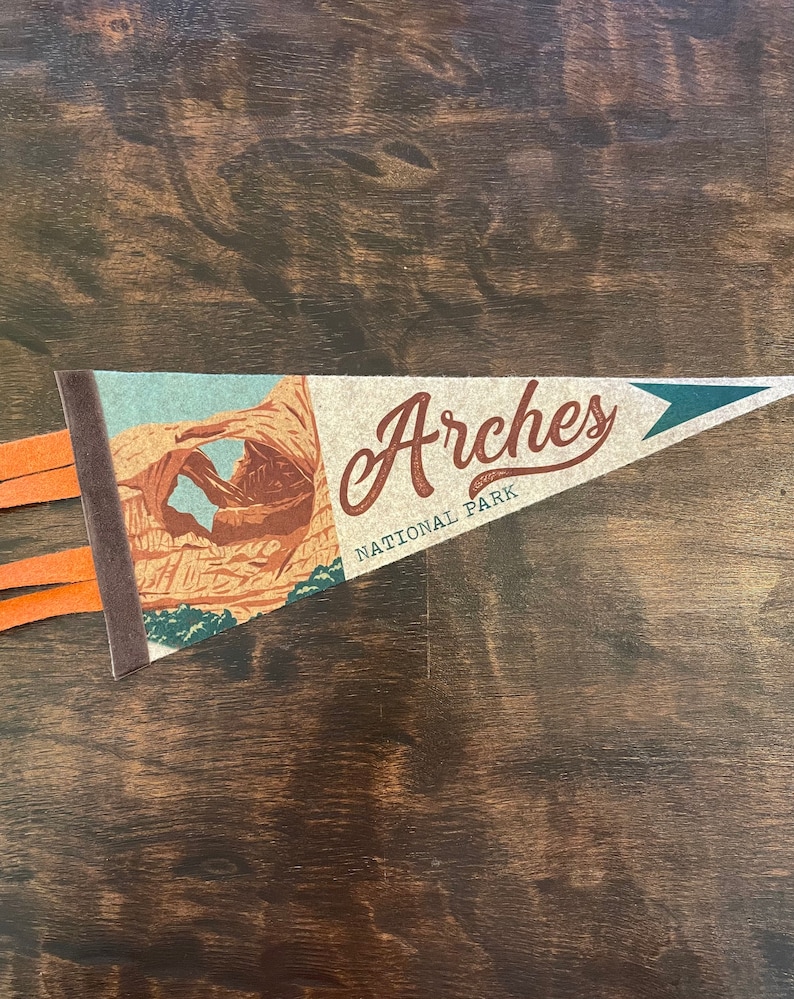 May include: A brown and tan pennant with a picture of a rock formation and the text "Arches National Park".