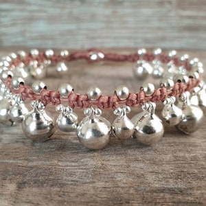 May include: A pink braided cord bracelet with silver bells hanging from it. The bracelet is on a wooden surface.