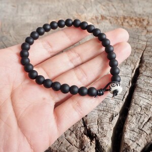 May include: A black beaded bracelet with a white bead with a black design. The bracelet is on a wooden surface.
