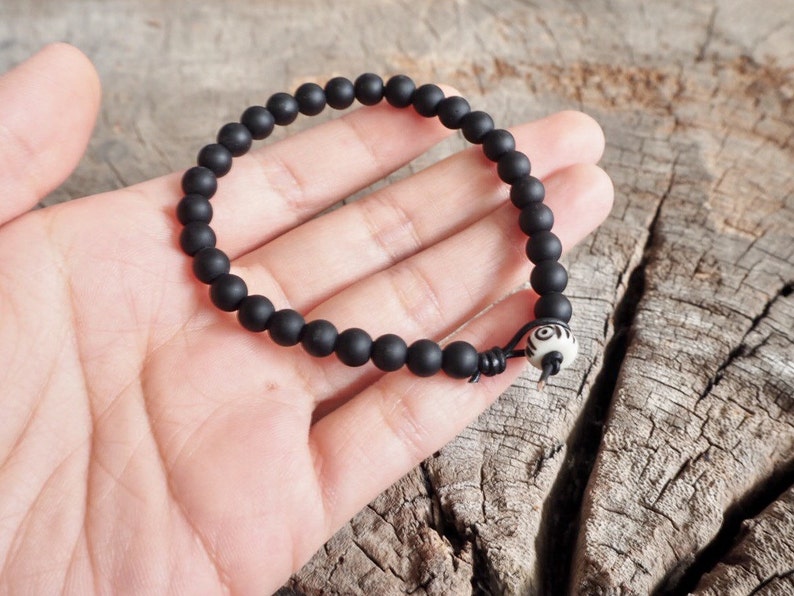 May include: A black beaded bracelet with a white bead with a black design. The bracelet is on a wooden surface.