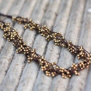 May include: A brown cord necklace with gold beads woven into a delicate pattern. The necklace is made of small, round beads that are clustered together to create a unique and intricate design.