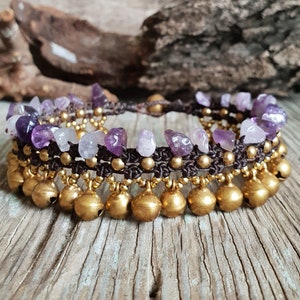 Puede incluir: Un tobillo morado y dorado con piedras de amatista y cascabeles dorados. El tobillo está hecho con cordón marrón y está atado con un nudo.