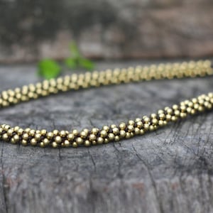 May include: A gold beaded necklace with a dark brown cord. The necklace is made of small, round beads that are strung together in a continuous pattern.