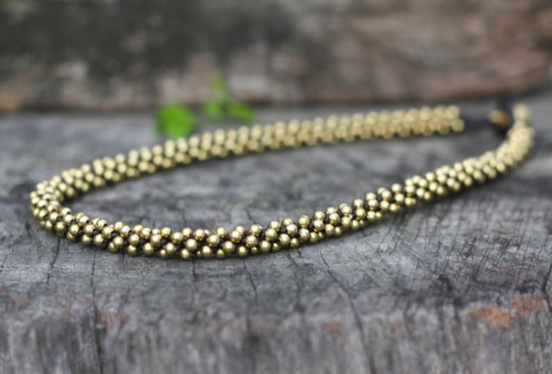 May include: A gold beaded necklace with a dark brown cord. The necklace is made of small, round beads that are strung together in a continuous pattern.