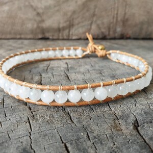 Puede incluir: Un brazalete de cuero blanco con cuentas de piedras preciosas blancas. El brazalete tiene un cierre dorado.