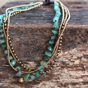 Puede incluir: Un collar de múltiples hilos con cuentas de piedras preciosas verdes y cuentas de metal dorado. El collar tiene un pequeño charm de campana al final.