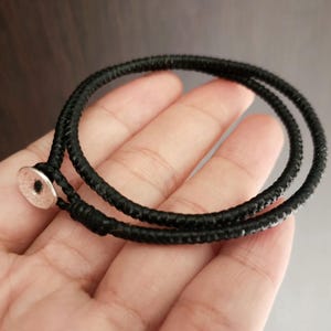 May include: A black bracelet with a woven texture, held in a hand. The bracelet wraps around twice and fastens with a small, silver-colored button clasp. The background is a blurred, dark brown.