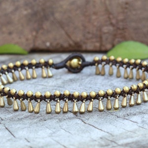 K&ouml;nnte beinhalten: Ein goldfarbenes Fu&szlig;kettchen mit brauner Kordelbasis. Das Fu&szlig;kettchen hat kleine, tropfenf&ouml;rmige Anh&auml;nger, die an der Kordel h&auml;ngen. Das Design umfasst kleine goldene Perlen. Das Fu&szlig;kettchen wird auf einer Holzoberfl&auml;che pr&auml;sentiert.