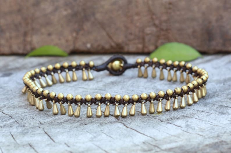K&ouml;nnte beinhalten: Ein goldfarbenes Fu&szlig;kettchen mit brauner Kordelbasis. Das Fu&szlig;kettchen hat kleine, tropfenf&ouml;rmige Anh&auml;nger, die an der Kordel h&auml;ngen. Das Design umfasst kleine goldene Perlen. Das Fu&szlig;kettchen wird auf einer Holzoberfl&auml;che pr&auml;sentiert.