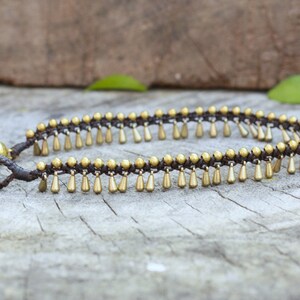 K&ouml;nnte beinhalten: Fu&szlig;kettchen mit einer dunkelbraunen Kordel und goldfarbenen Tropfenanh&auml;ngern. Das Armband hat eine kleine goldene Glocke und wird auf einer Holzoberfl&auml;che pr&auml;sentiert. Das Design ist schlicht und elegant.