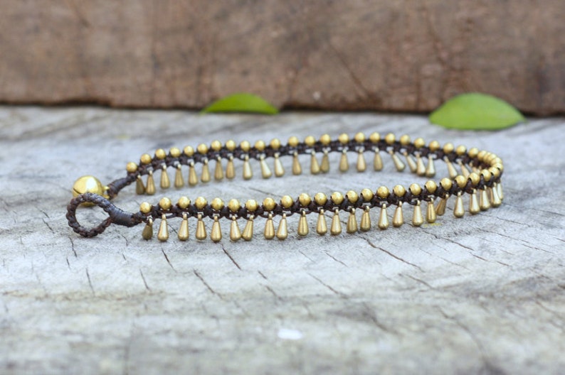 K&ouml;nnte beinhalten: Fu&szlig;kettchen mit einer dunkelbraunen Kordel und goldfarbenen Tropfenanh&auml;ngern. Das Armband hat eine kleine goldene Glocke und wird auf einer Holzoberfl&auml;che pr&auml;sentiert. Das Design ist schlicht und elegant.