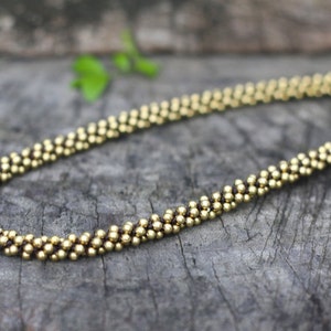 May include: A gold beaded necklace with a brown cord and a small gold bead at the end. The necklace is lying on a wooden surface.
