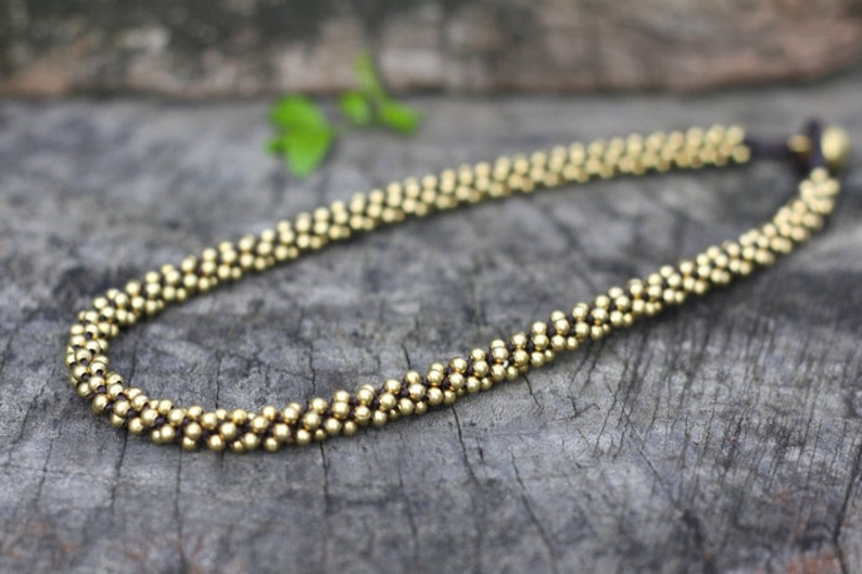May include: A gold beaded necklace with a brown cord and a small gold bead at the end. The necklace is lying on a wooden surface.