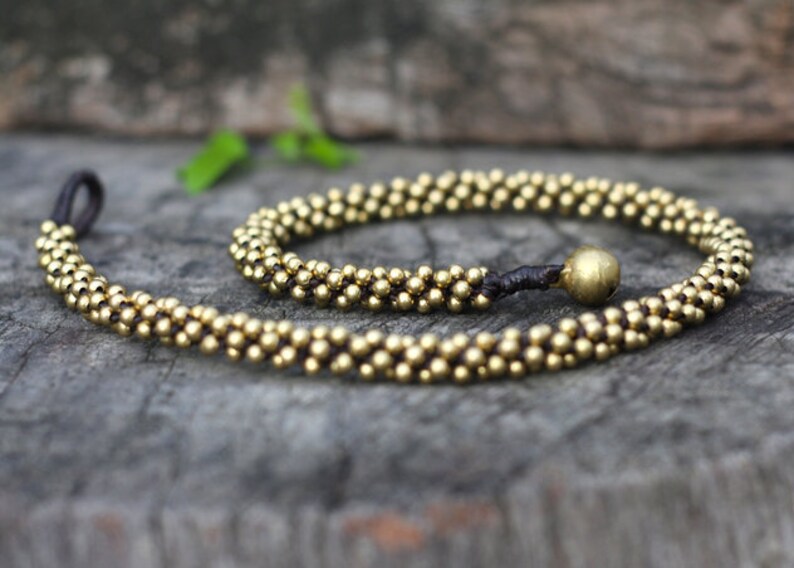 May include: A gold beaded necklace with a dark brown cord and a gold bead accent. The necklace is made of small, round beads and is approximately 18 inches long.