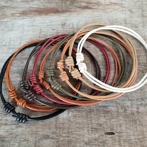 Könnte beinhalten: Ein Set aus acht Lederarmbändern mit geknüpften Verschlüssen in verschiedenen Farben, darunter Schwarz, Braun, Rot, Grün und Weiß.