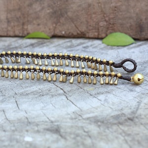 K&ouml;nnte beinhalten: Ein Fu&szlig;kettchen mit braunem Kordel und goldfarbenen Perlen und tropfenf&ouml;rmigen Anh&auml;ngern. An einem Ende h&auml;ngt eine kleine goldene Glocke. Der Schmuck wird auf einer verwitterten Holzoberfl&auml;che pr&auml;sentiert.