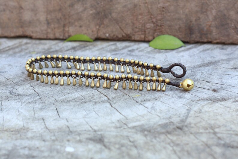 K&ouml;nnte beinhalten: Ein Fu&szlig;kettchen mit braunem Kordel und goldfarbenen Perlen und tropfenf&ouml;rmigen Anh&auml;ngern. An einem Ende h&auml;ngt eine kleine goldene Glocke. Der Schmuck wird auf einer verwitterten Holzoberfl&auml;che pr&auml;sentiert.