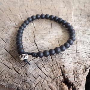 May include: A black beaded bracelet with a white bone bead with a geometric pattern. The bracelet is on a wooden surface.