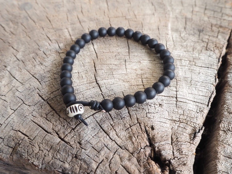 May include: A black beaded bracelet with a white bone bead with a geometric pattern. The bracelet is on a wooden surface.