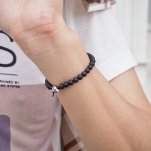 May include: A black beaded bracelet with a silver charm. The bracelet is worn on a person's wrist.