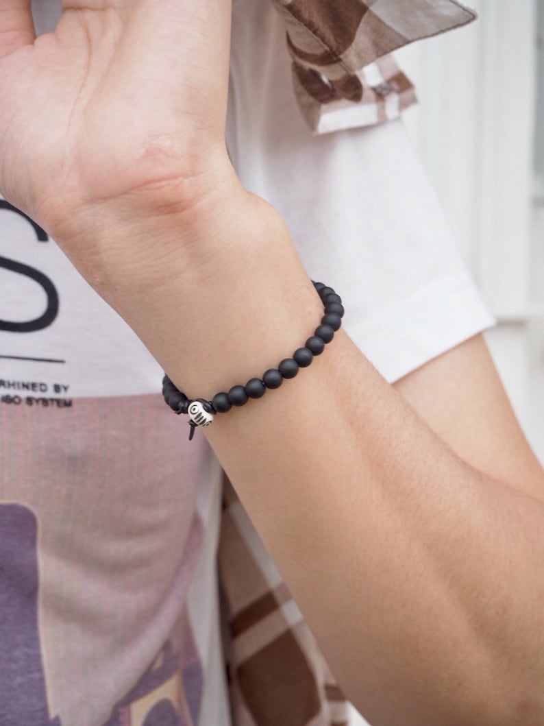 May include: A black beaded bracelet with a silver charm. The bracelet is worn on a person's wrist.