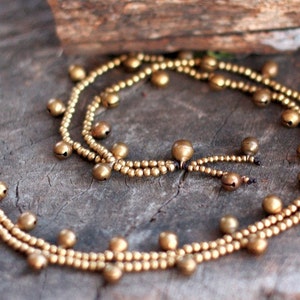 May include: A gold anklet with small bells, made of two strands of beads. The anklet is lying on a wooden surface.