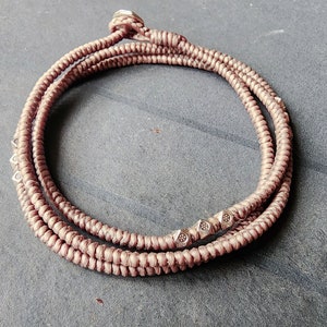 Könnte beinhalten: Ein braunes Kordel-Armband mit silbernen Perlen. Das Armband ist mehrmals um sich selbst gewickelt.