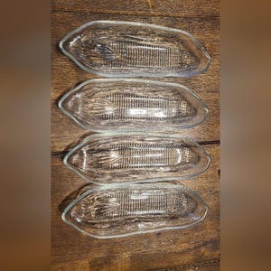 May include: Four clear glass corn-shaped dishes. Each dish has a textured interior, mimicking the appearance of corn on the cob. The dishes are arranged on a wooden surface, showcasing their design and shape.