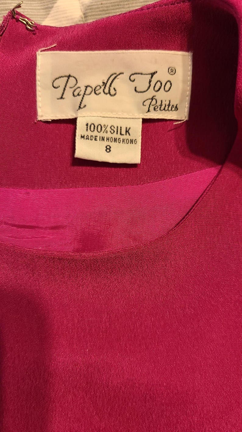 Peut inclure: Une &eacute;tiquette blanche avec du texte noir qui indique "Papell Too Petites" et "100% Soie Fabriqu&eacute; &agrave; Hong Kong 8". L'&eacute;tiquette est attach&eacute;e &agrave; un tissu de soie fuchsia.