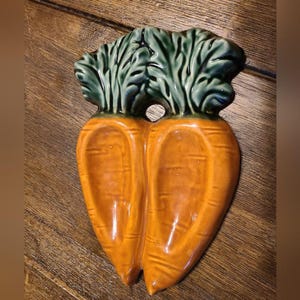 May include: A ceramic dish shaped like two joined carrots with a glossy orange finish and textured green leafy tops. The piece features a small circular hole between the carrot tops, a smooth surface, and distinct horizontal ridges along the orange bodies.