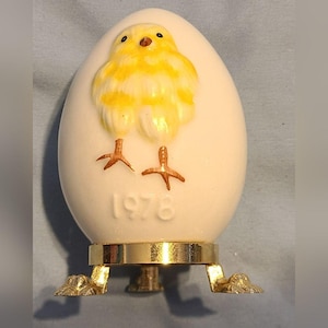 Puede incluir: Figura de cerámica blanca en forma de huevo con un diseño de pollito amarillo y el año 1976. Se apoya en un soporte de metal dorado con tres patas. El pollito tiene plumas amarillas y blancas y patas marrones. Un artículo decorativo de Pascua.