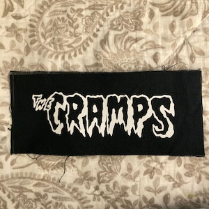 May include: Black rectangular patch with the white text "THE CRAMPS". The text has a dripping, stylized font. The patch is on a patterned, neutral-toned fabric.