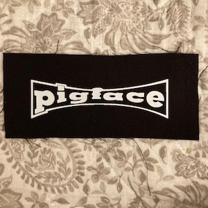 Puede incluir: Un parche rectangular negro con la palabra "pigface" en blanco en una fuente estilizada. El parche tiene un borde blanco y está sobre un fondo estampado. Los bordes del parche tienen hilos sueltos.