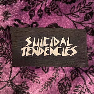 Puede incluir: Un parche rectangular de tela negra con el texto blanco Suicidal Tendencies en una fuente angular estilizada. El parche tiene una textura tejida y está colocado sobre un fondo morado con un patrón floral oscuro e intrincado.