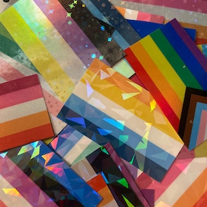Puede incluir: Una colección de pegatinas rectangulares con varios diseños de banderas del orgullo con acabados holográficos. Las pegatinas muestran vibrantes rayas horizontales, patrones geométricos y motivos de estrellas en un espectro de colores.
