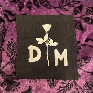 Può includere: Una toppa quadrata in tessuto nero con una stampa grafica bianca di un singolo stelo di rosa posizionato tra le lettere maiuscole D e M. Il design è centrato sulla superficie tessile scura, mostrando un netto contrasto tra l'inchiostro bianco e il materiale di fondo nero.