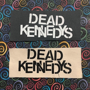Op de afbeelding: Twee stoffen patches met de tekst "DEAD KENNEDYS" in een stencil lettertype. De bovenste patch is zwart met witte tekst. De onderste patch is beige met zwarte tekst.