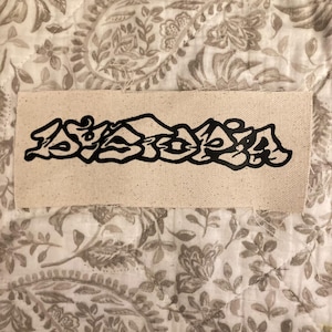 Peut inclure: Un patch rectangulaire en tissu beige avec des lettres noires de style graffiti. Le patch est rectangulaire et semble être fait de toile ou d'un matériau similaire. L'écriture noire est stylisée et abstraite.