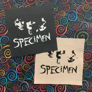 May include: Two iron-on patches with the word "SPECIMEN" in white lettering on a black patch and in black lettering on a beige patch. Both patches feature a stylized image of a skull with two eyes.