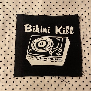 May include: A square black fabric patch featuring a white screen-printed graphic of a record player and the text Bikini Kill in a stylized font. The patch has raw, slightly frayed edges and a visible woven texture on the black fabric surface.