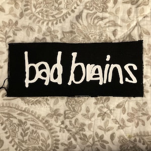 May include: A rectangular black patch with the words "bad brains" in white, stylized lettering. The patch is likely made of fabric and has a stitched border. The background is a patterned fabric.